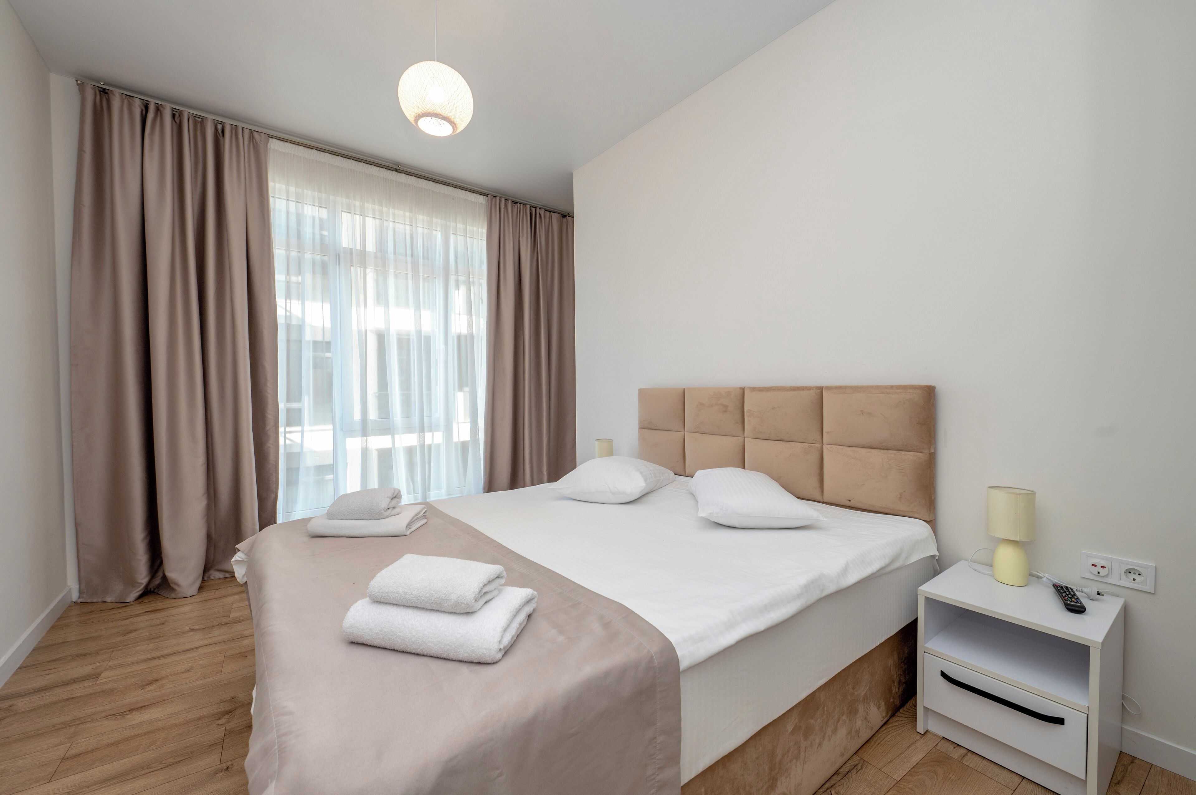 A neatly made bed with white linens and a beige headboard, a bedside table with a lamp, and a window with sheer curtains.