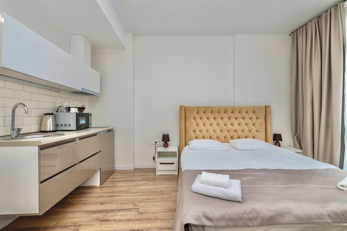 Habitación de hotel moderna con kitchenette, cama con cabecera capitonada y ropa de cama ordenada con toallas.