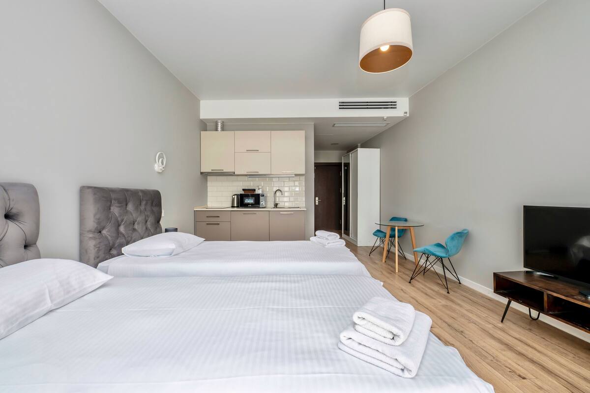 Una habitación de hotel moderna con una cama grande, televisión, un pequeño espacio con cocina y una zona de estar con sillas.