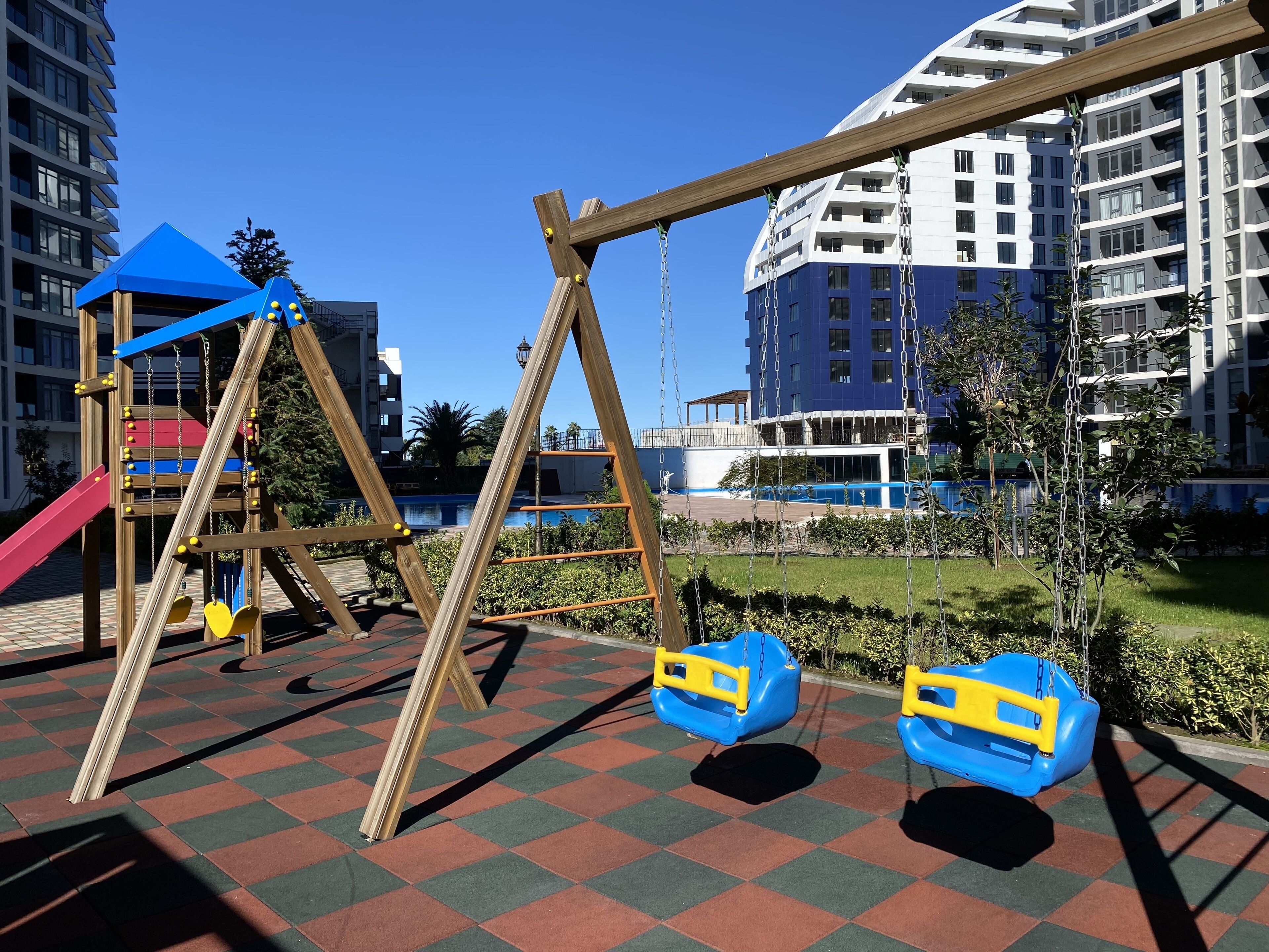 A playground with swings and a slide, surrounded by a checkered floor and tall residential buildings in the background.
