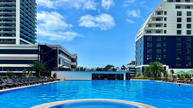 Una gran piscina al aire libre con un jacuzzi en el centro, rodeada de tumbonas y edificios modernos al fondo.