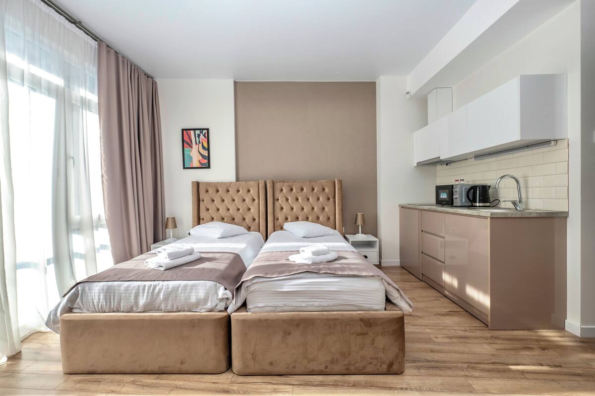 Una cama doble con cabecera, una mesita de noche, una pequeña kitchenette y una ventana con cortinas.