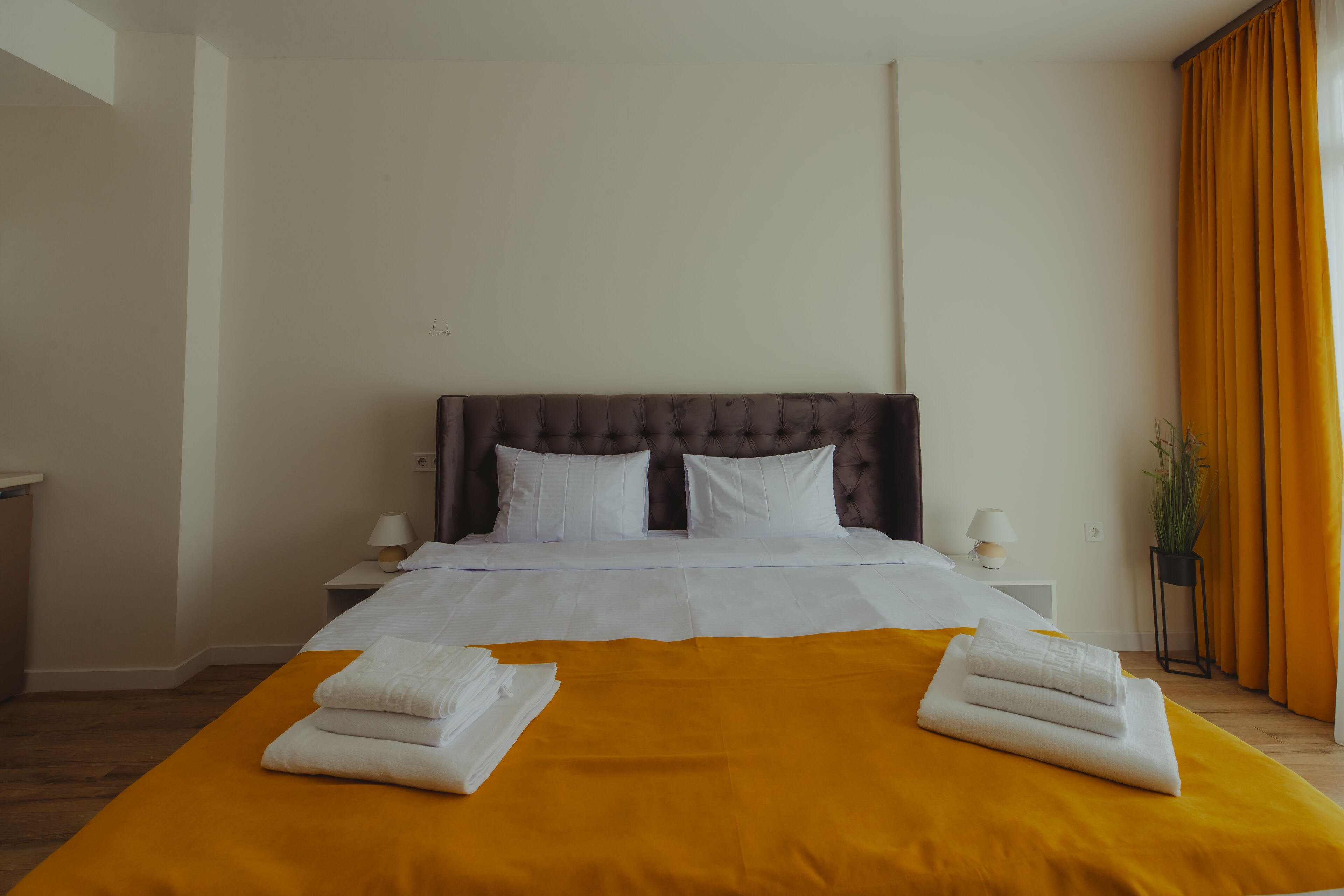 Een net opgemaakt bed met witte lakens en een gele spreidek, twee witte handdoeken op het bed, een donker hoofdbord, en een plant in een pot naast het raam.