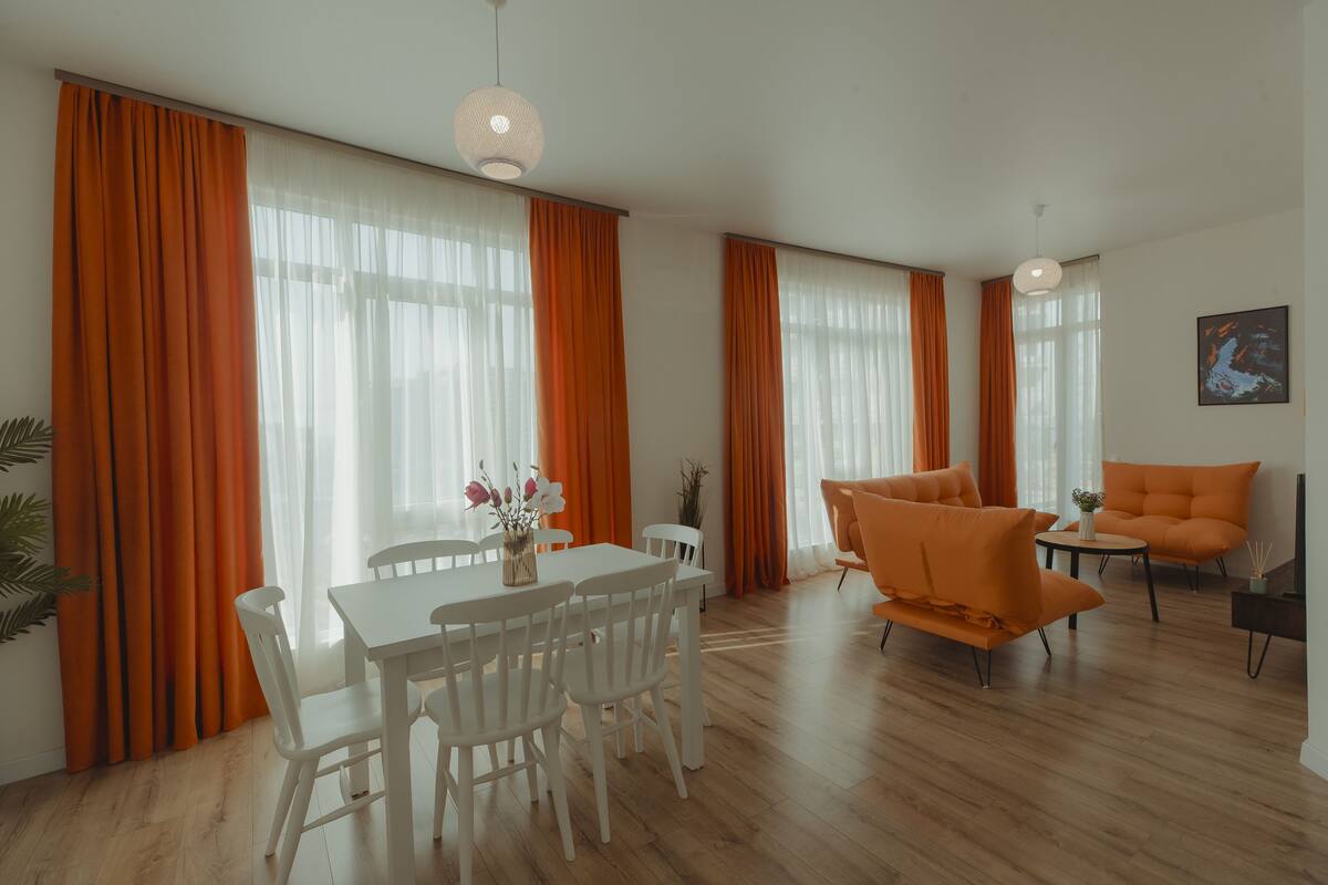 Un amplio espacio de sala con una mesa de comedor, sillas blancas, sofás naranja y ventanas grandes con cortinas naranja.