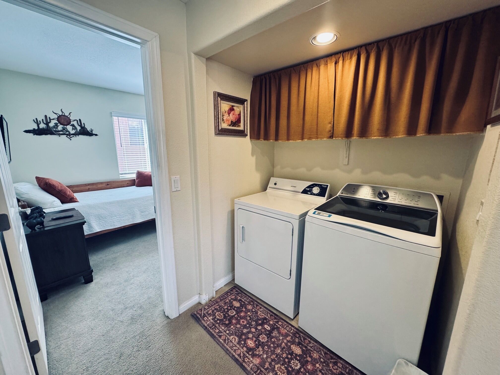 A compact laundry room with a washing machine and dryer, a small rug, and a bed in the adjacent room.