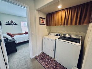 A compact laundry room with a washing machine and dryer, a small rug, and a bed in the adjacent room. - Fox Run | 3BR Near Wolf Creek + Free Activities (Mesquite)