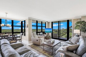 A spacious living room with a large window offering a view of the ocean, a comfortable sofa, a dining area, and a modern chandelier.