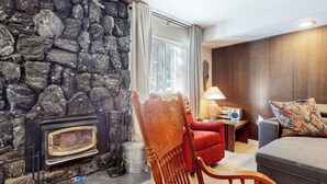 A cozy living room with a stone wall, a fireplace, a red armchair, a grey sofa, and a wooden panel wall. - Central Location, Restaurants & Shops. Tennis Courts, Hot Tub & Pool On-site (Mammoth Lakes)