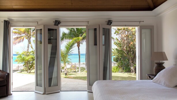 Une chambre avec un grand lit, une table de chevet et une lampe. De grandes portes coulissantes en verre donnent sur un balcon d’où l’on peut admirer la plage et les palmiers.