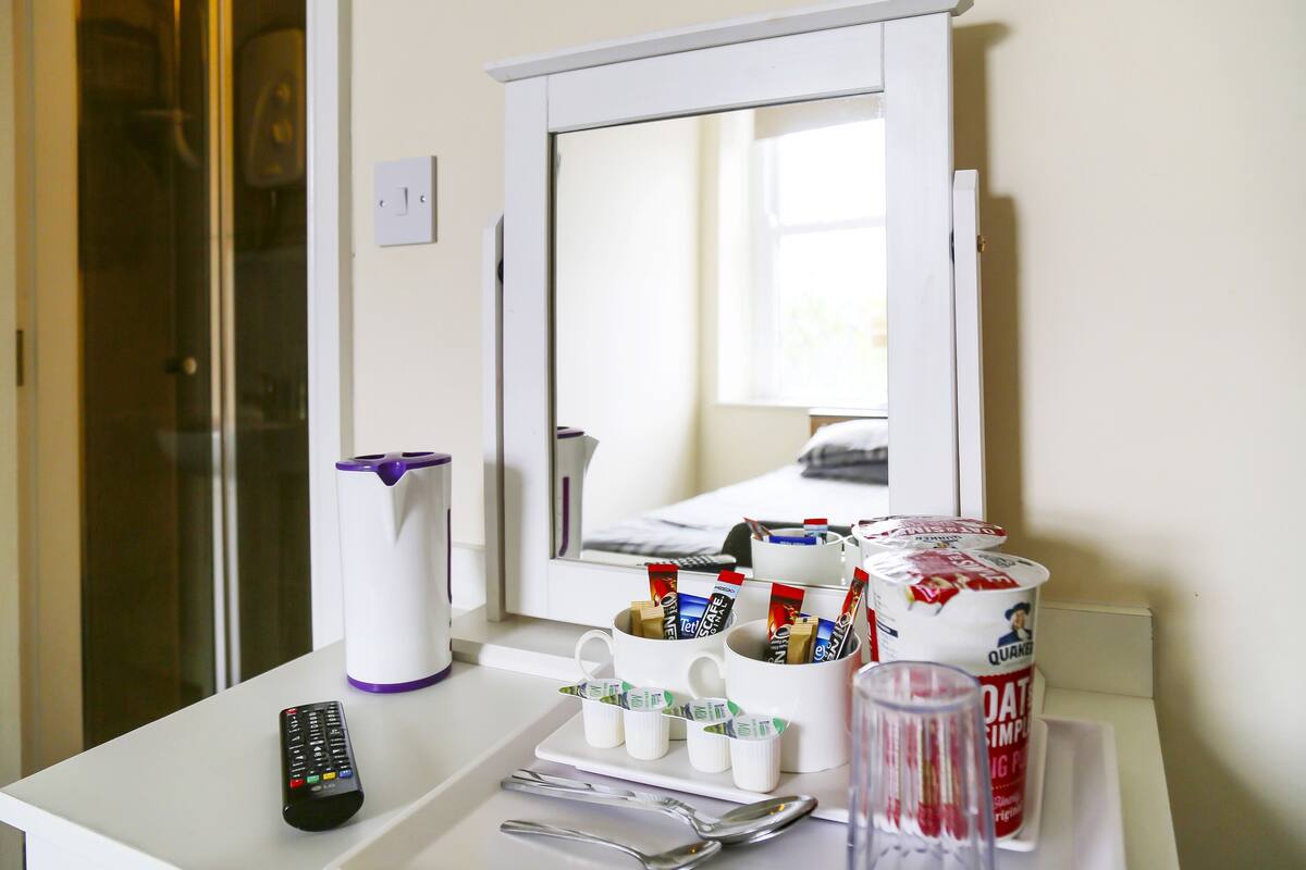 A hotel room with a white bedside table, a remote control, a coffee maker, a glass, and a container of Quaker Oats cereal.