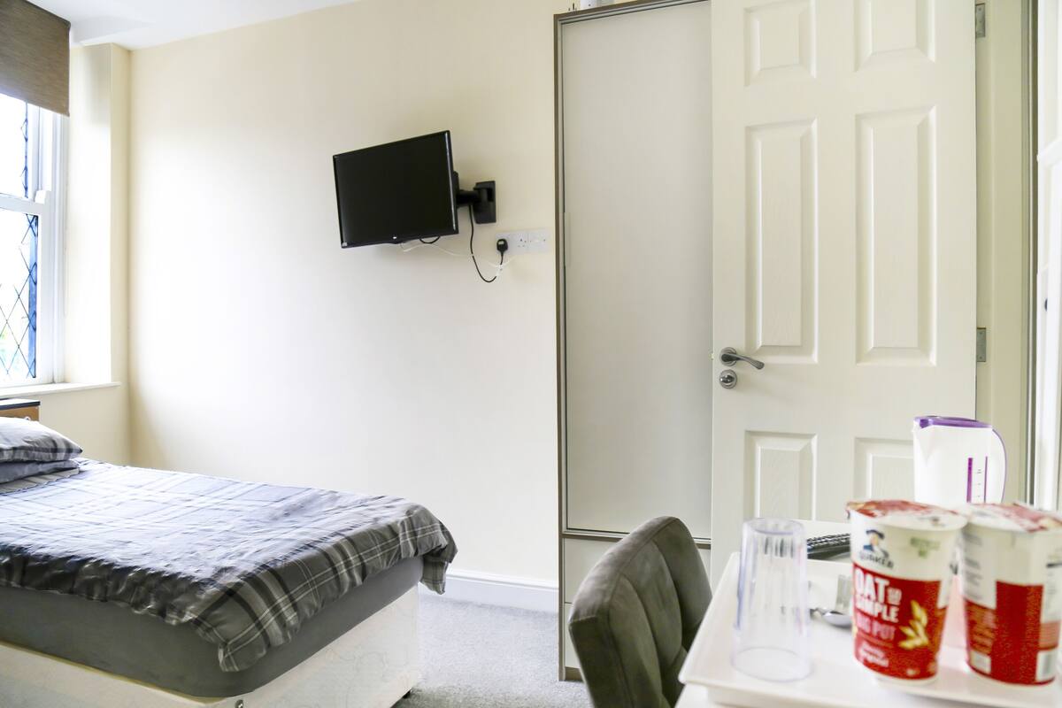 A bedroom with a bed, a television mounted on the wall, a door, and a table with a can of food and a glass.