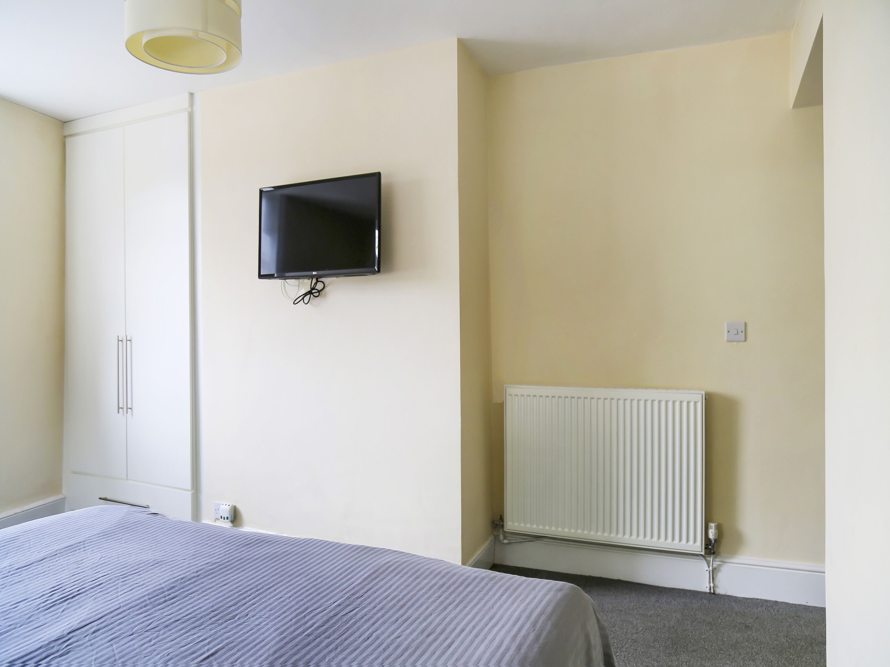 A bedroom with a bed, a wardrobe, a television mounted on the wall, and a radiator.