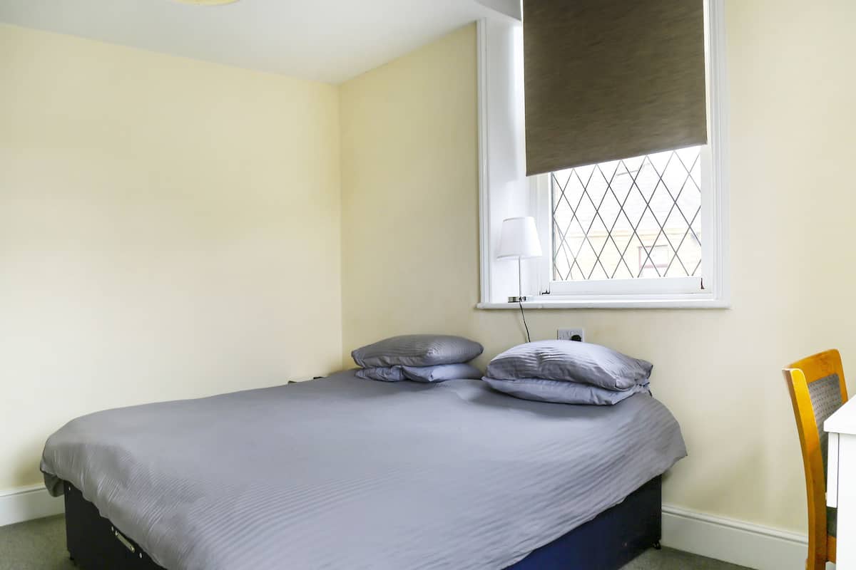 A single bed with a grey mattress and pillows, a bedside table with a lamp, and a window with a metal grid.