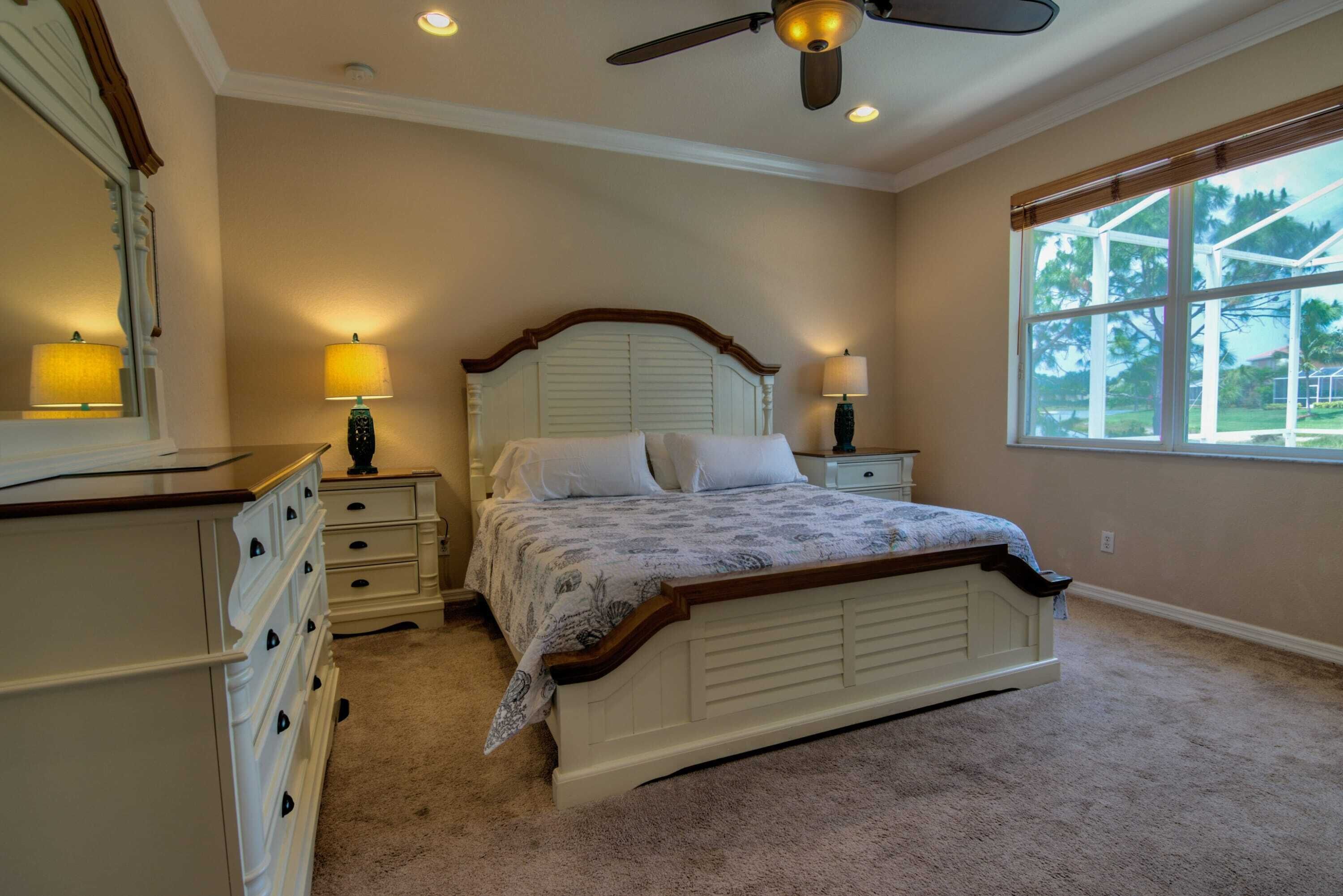 A bedroom with a bed, nightstands, a dresser, a ceiling fan, and a window with a view.