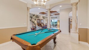 A pool table in a well-lit room with a chandelier, a sofa, and a view of the outdoors. - Lakeside Living, Private Beach, Pool Table, Two Heated Pools - The Joint Venture - Roelens Vacations (Cape Coral)