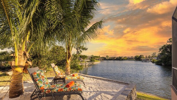 A serene riverside patio with lounge chairs and a palm tree, overlooking a calm river and a sunset sky. - Lakeside Living, Private Beach, Pool Table, Two Heated Pools - The Joint Venture - Roelens Vacations (Cape Coral)