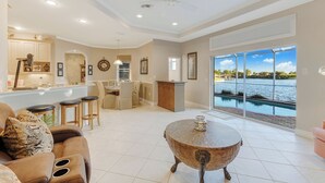 A spacious living area with a dining set, a round table with a candle, and a large window overlooking a body of water. - Lakeside Living, Private Beach, Pool Table, Two Heated Pools - The Joint Venture - Roelens Vacations (Cape Coral)