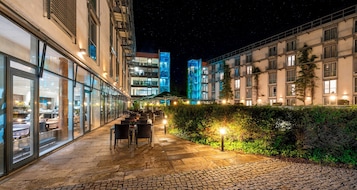 Un patio exterior iluminado de noche con mesas y sillas, setos y edificios con ventanas y balcones iluminados.