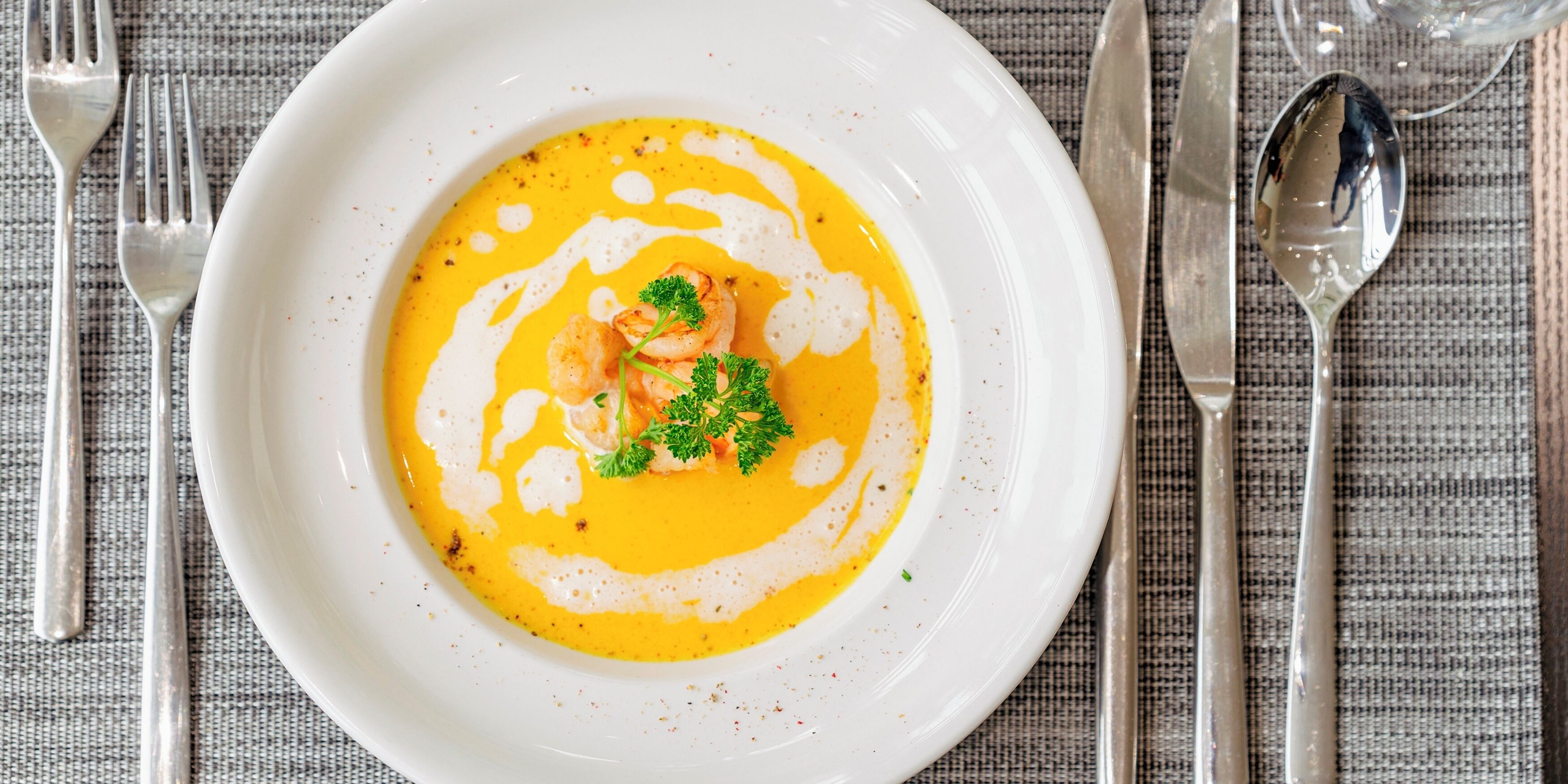 A white plate with a creamy soup garnished with parsley and shrimp, accompanied by a spoon and fork on a textured tablecloth.