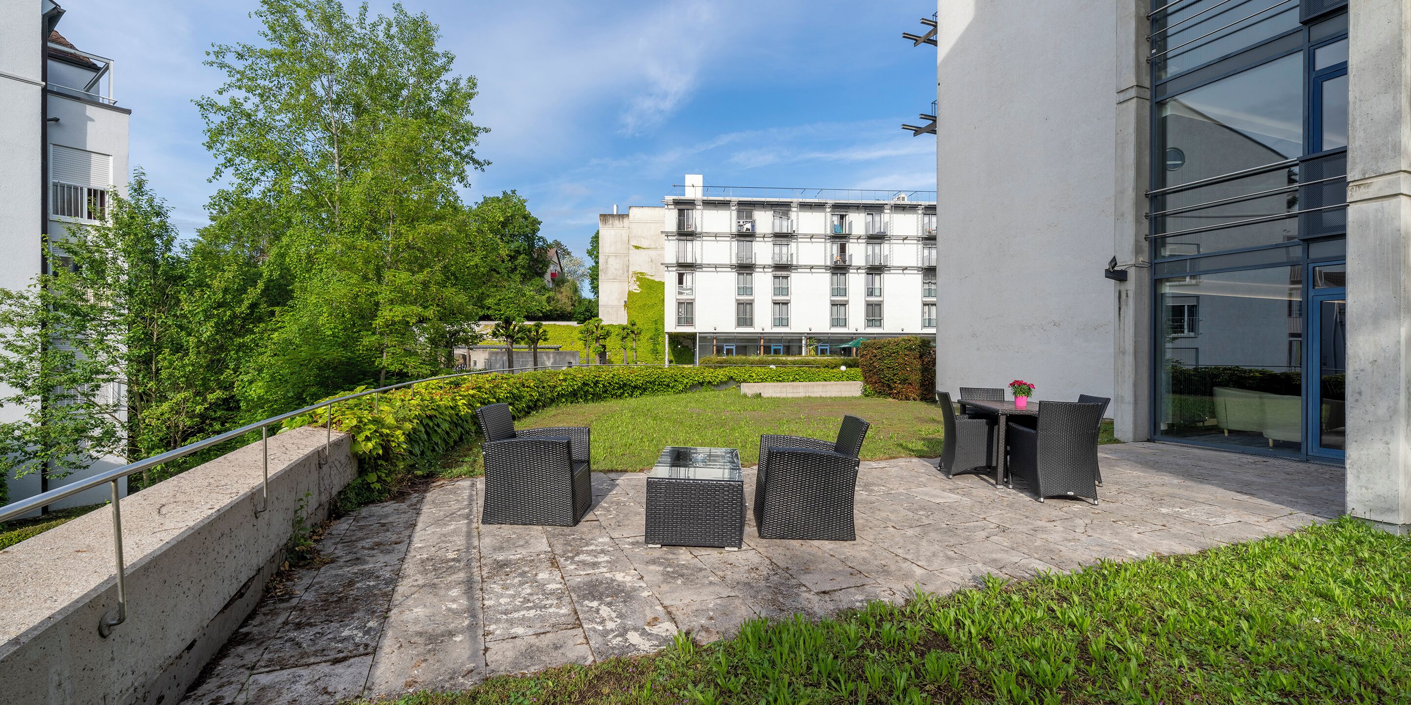 Un patio al aire libre con mobiliario de mimbre, un camino de hormigón y vistas a un edificio con varias ventanas.