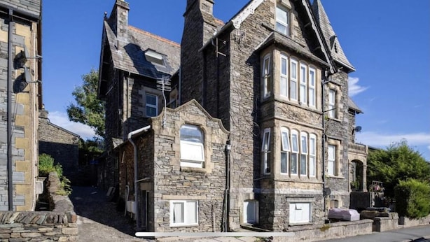 A stone building with multiple chimneys, large windows, and a steeply pitched roof. - The Mews, With Free Private Parking, Windermere (Windermere)