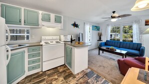A compact kitchen with white and teal cabinets, a white stove, and a microwave. There's a blue sofa and a red chair in the living area, with a ceiling fan and a window with curtains. - Lake Placid Home Boat Parking and Clubhouse Access by RedAwning (Lake Placid)