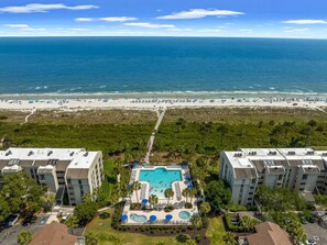 A resort with a central swimming pool surrounded by lounge chairs and umbrellas, overlooking a beach and ocean.