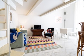 A living room with a sofa, a TV, a wooden cabinet, a colorful rug, and a dining area with chairs.