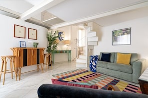 A living room with a sofa, a coffee table, a patterned rug, a staircase, and a dining area with a bar.