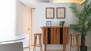 A room with a round table, two bar stools, a central rattan table, framed pictures on the wall, a cactus, and a large potted plant.