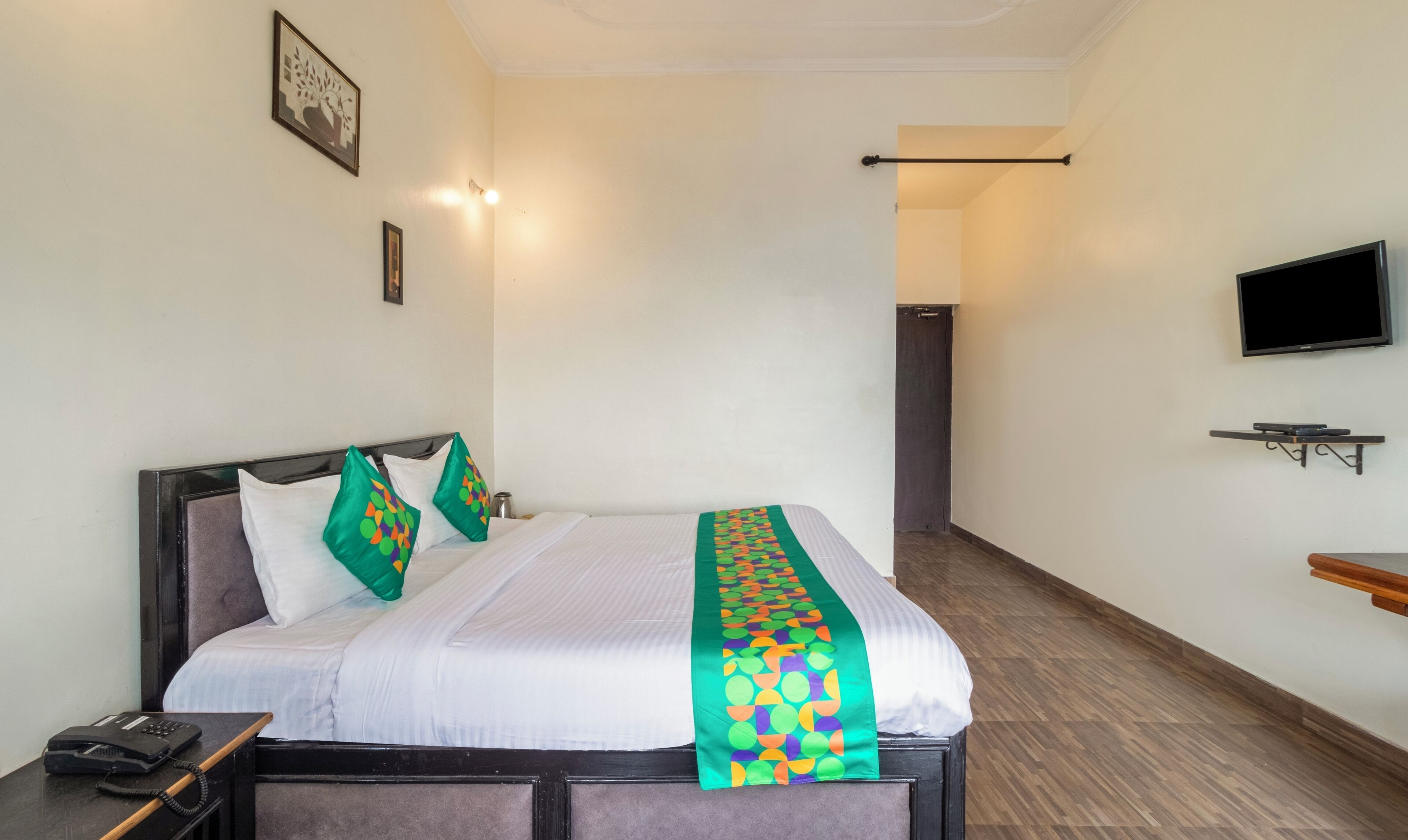 A hotel room with a bed, a bedside table with a phone, a TV mounted on the wall, and a door leading to another room.