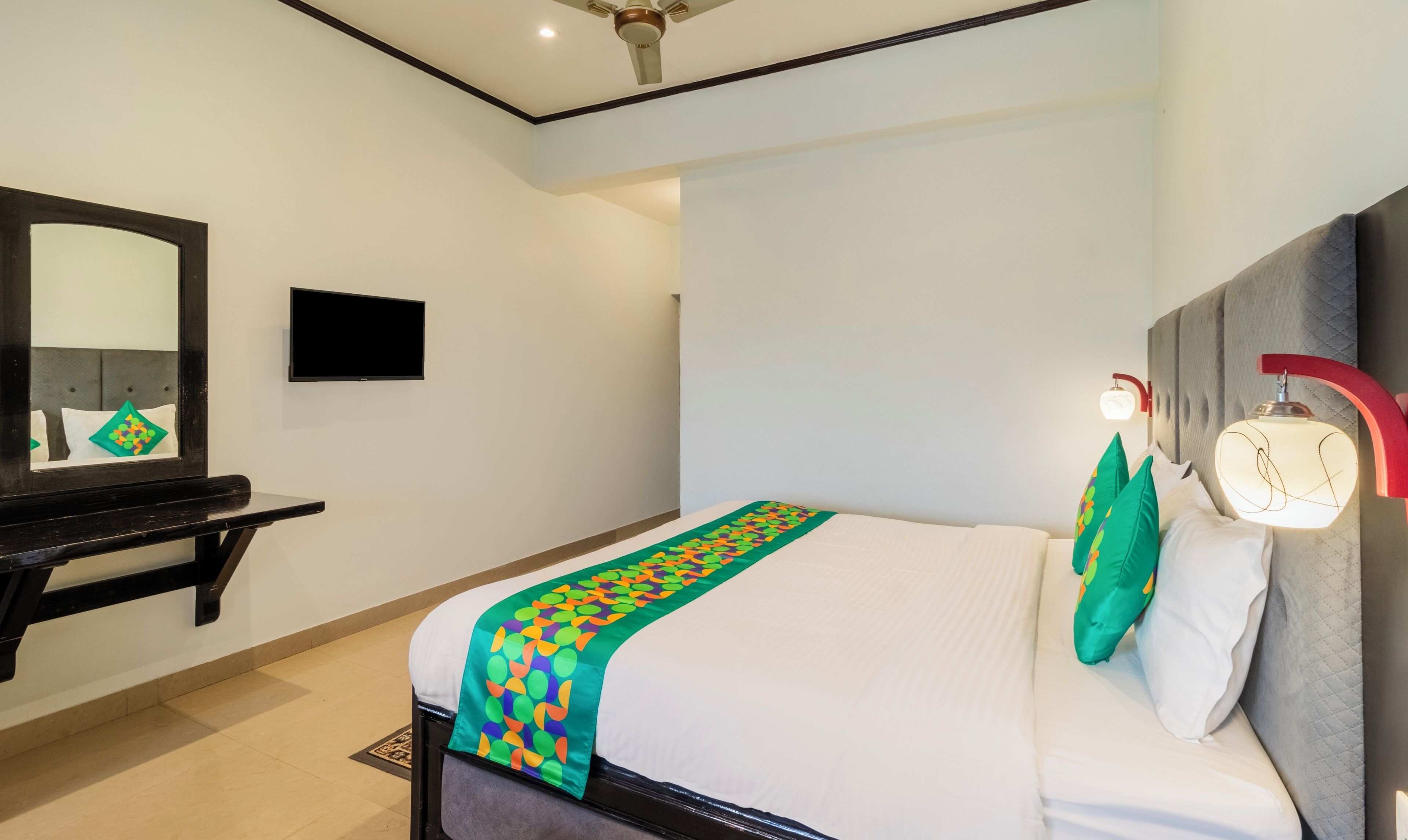 A hotel room with a bed, a bedside table, a mirror, a wall-mounted TV, and a ceiling fan.