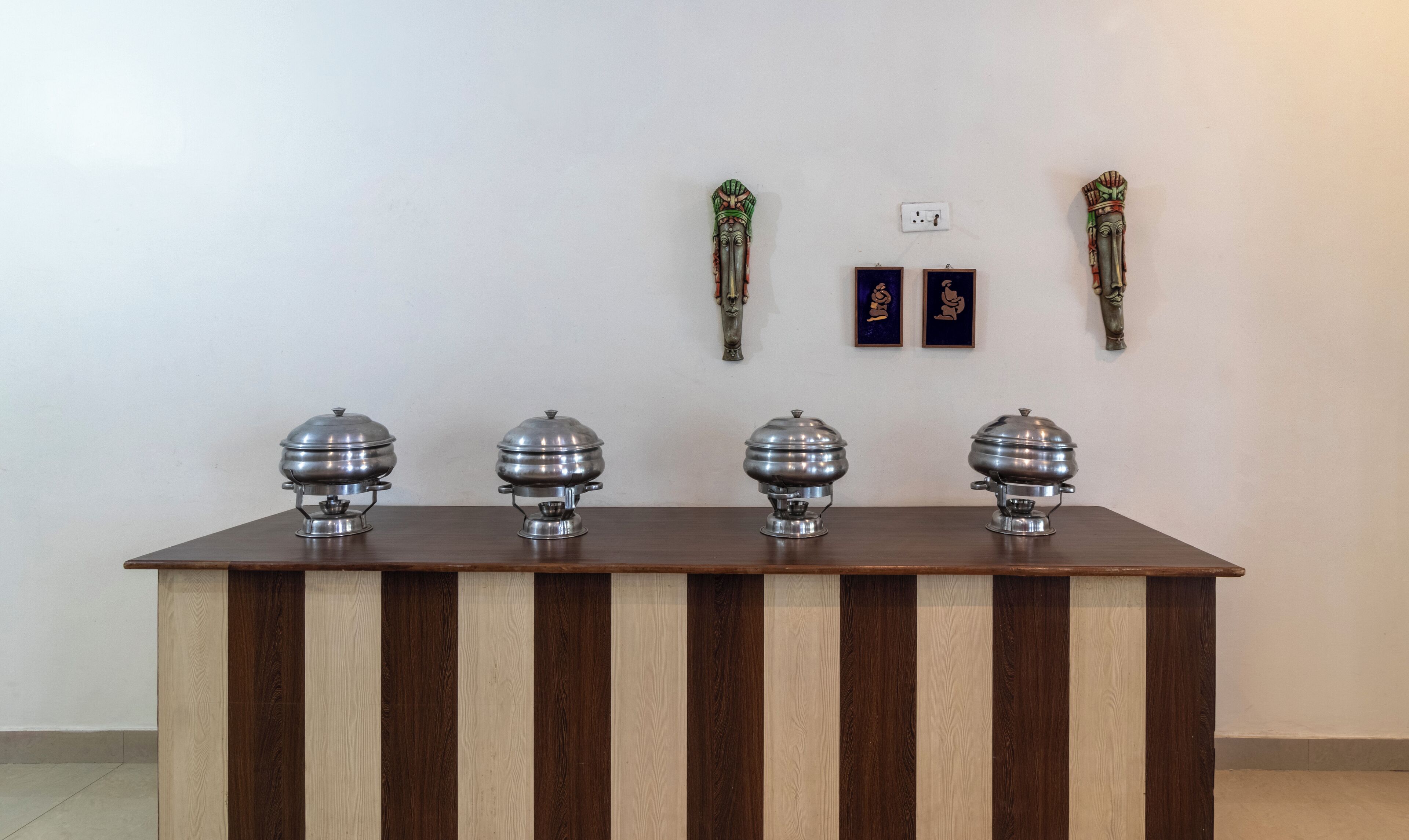 Una mesa de madera con cuatro objetos plateados, posiblemente recipientes de comida, frente a una pared lisa con dos cuadros enmarcados y un enchufe.