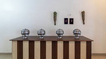 Una mesa de madera con cuatro objetos plateados, posiblemente recipientes de comida, frente a una pared lisa con dos cuadros enmarcados y un enchufe.