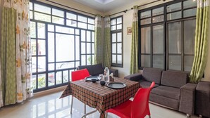 A room with a dining table set for two, a sofa, and large windows with curtains. - Treebo Spring Birds - 3Kms from Bhimtal Lake (Nainital)