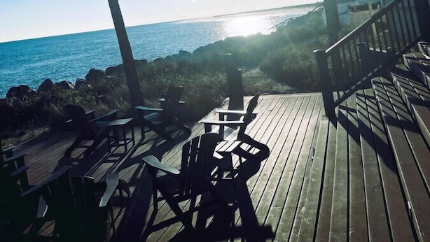En træterrasse med udendørs møbler, der giver udsigt over en strand med palmer og en bygning i baggrunden.