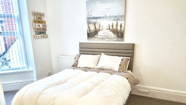 A neatly made bed with a grey headboard, a window with blinds, and a beach-themed painting on the wall. - Studio for 2 (ground floor) in fashionable North Shore (Blackpool)