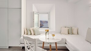 A modern, minimalist room with a white sofa, a small round table, and a view of the sea through an open window. - Santorini Mansion Maisonette Sea View Jacuzzi 3BDR (Thira - Santorini)