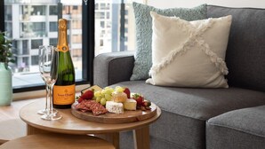 Quarto com um sofá cinzento, uma mesa de madeira com uma garrafa de Veuve Clicquot champagne, copos e uma tábua de queijos com diferentes queijos e frutas.