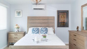 A bedroom with a wooden bed, bedside tables, a nightstand, a wall-mounted air conditioner, and a framed picture. - 1BR/1BA New Oceanside Condo (Charlotte Amalie)