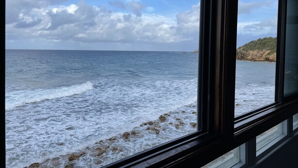 A view of the sea through a window with a rocky shoreline and a distant hill. - 1BR/1BA New Oceanside Condo (Charlotte Amalie)
