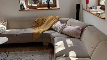 Ein modernes Wohnzimmer mit einem beigefarbenen Ecksofa, einem gemusterten Teppich, einem Holzfensterbrett mit Büchern und einer Vase sowie einer Stehlampe.