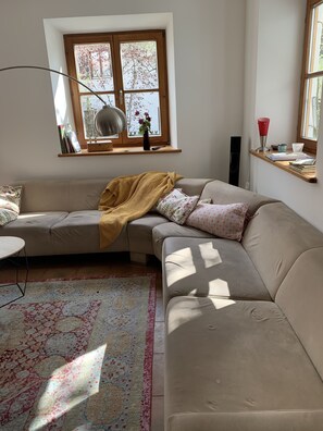 Uma sala de estar moderna com um sofá seccional bege, um tapete estampado, um peitoril de janela de madeira com livros e um vaso, e um abajur.