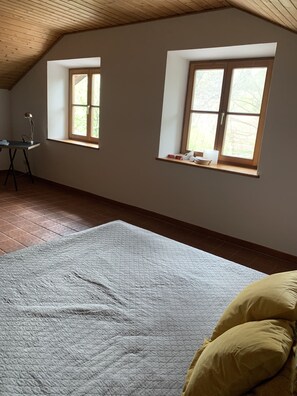 Um quarto com teto inclinado, duas janelas com molduras de madeira, uma cama com colcha e uma escrivaninha com luminária.
