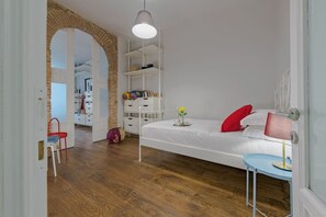 Quarto com cama branca, travesseiro vermelho, mesa de cabeceira com abajur e piso de madeira.