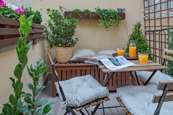 Un espace de détente extérieur accueillant, avec une table en bois, des chaises rembourrées, un pot de fleurs en osier et un magazine.
