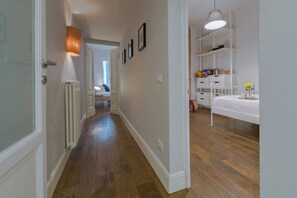 Un couloir doté d’un plancher en bois, de murs blancs, d’un radiateur, d’un lit à baldaquin blanc et d’une étagère blanche contenant divers objets.