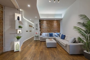 A modern living room with a curved white wall, a wooden floor, a grey sectional sofa with blue cushions, a small side table, and a potted plant.