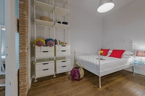 Et soveværelse med en hvid seng, røde puder, et hvidt metal sengegavl, en hvid kommode med skuffer, en fletkurv og en hængende lampe.