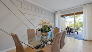 Ein Essbereich mit Glastisch und Stühlen, einer Vase mit weißen Blumen und Blick ins Freie durch große Fenster.